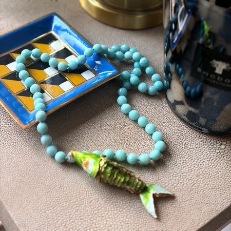 Turquoise beaded necklace with a fish pendant on a textured surface.